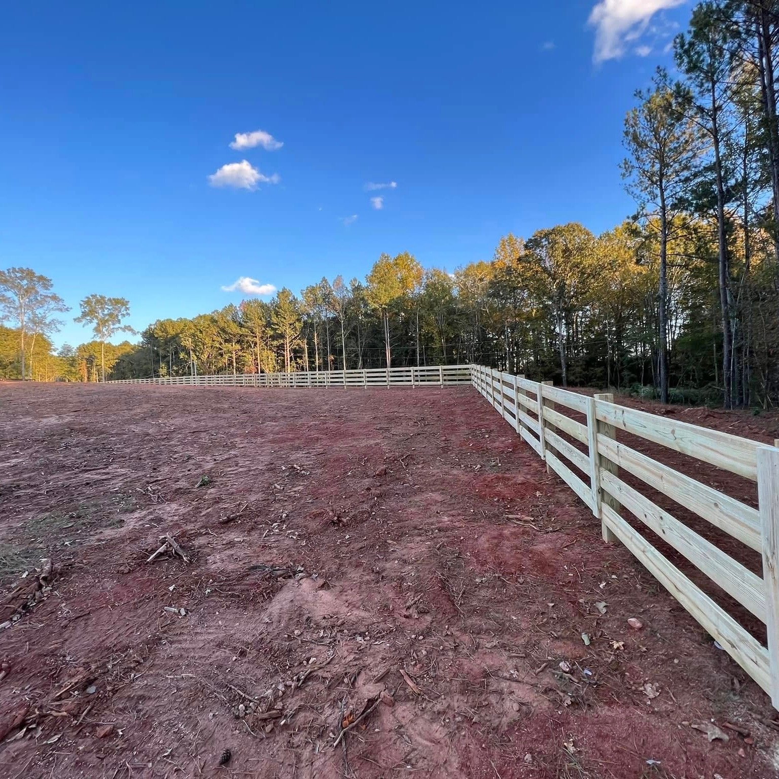 A dirt field with a wooden fence along the right side, with trees in the background and a clear blue sky with a few clouds.