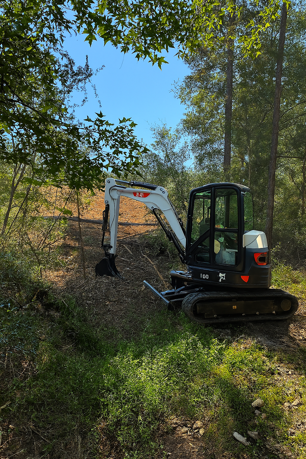 Small black and white excavator with a closed cabin, working on a hillside in a forested area under a blue sky.