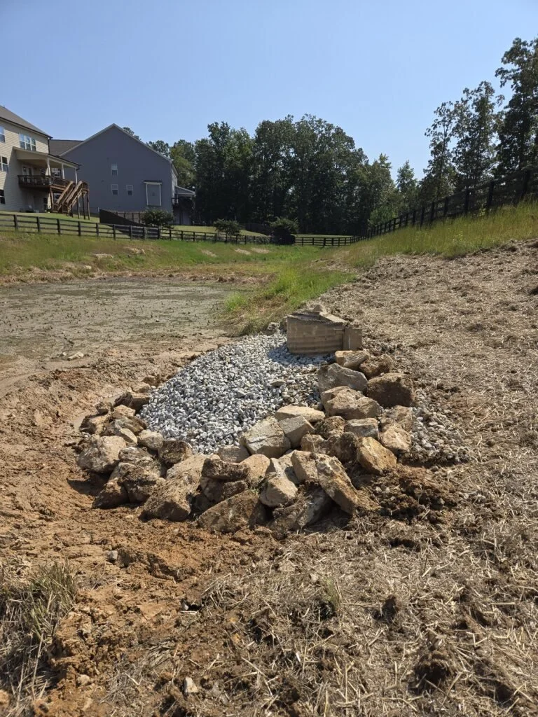 A landscape view of a sloped backyard with a drainage ditch under construction filled with gravel and surrounded by rocks, with houses and trees in the background.