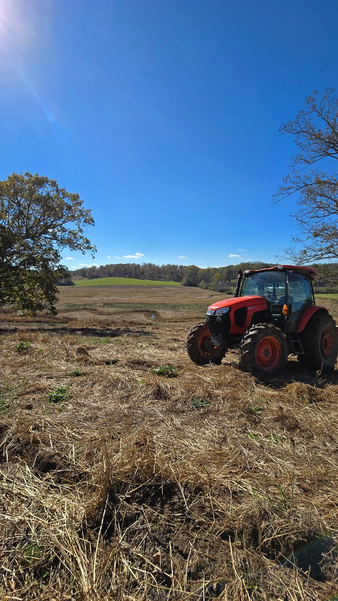A red tractor parked on a field with dry grass, trees on the left and right, and a blue sky overhead.