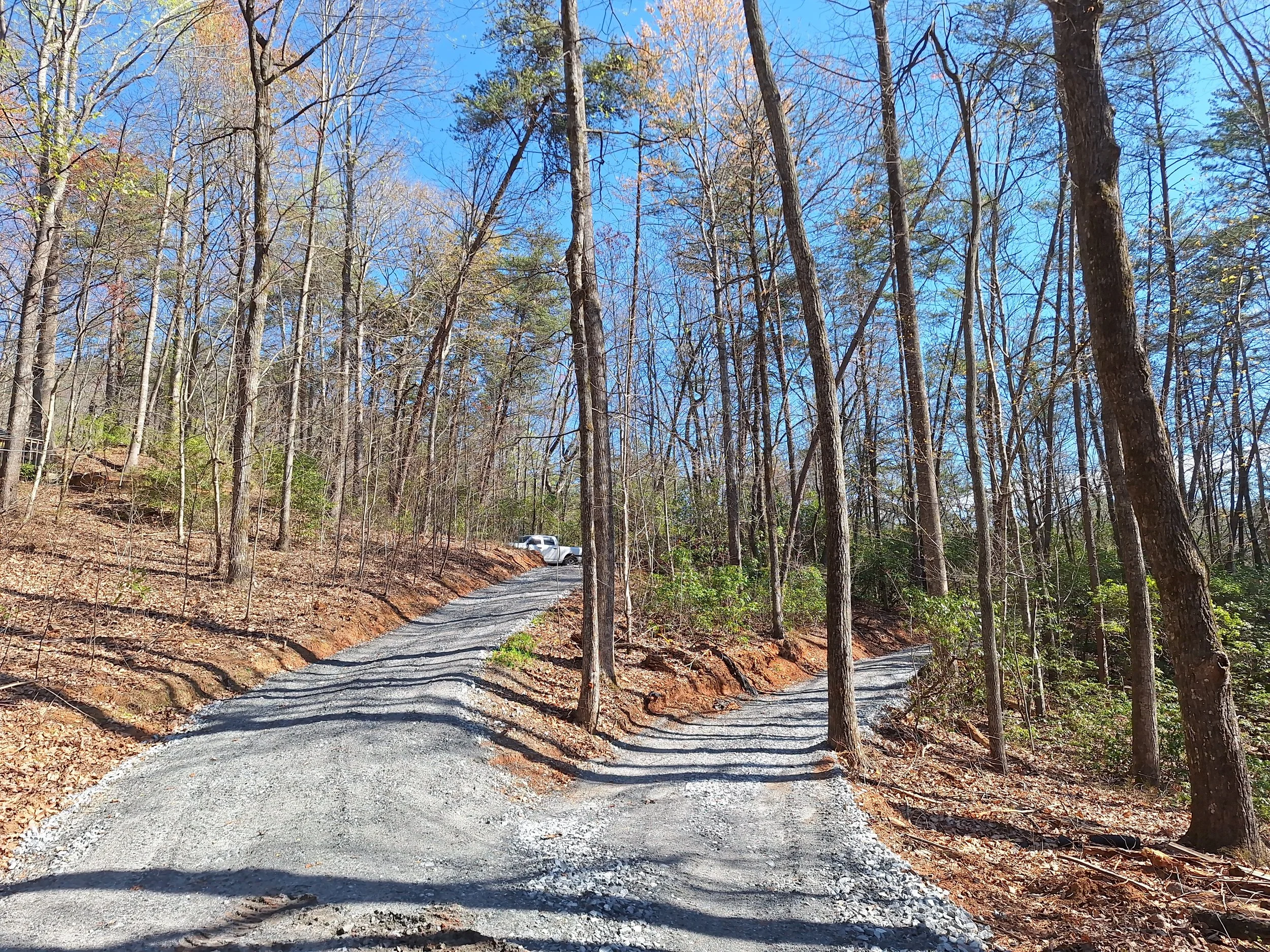 A gravel road winding through a wooded forest on a sunny day, with a white vehicle parked near the top of the hill.