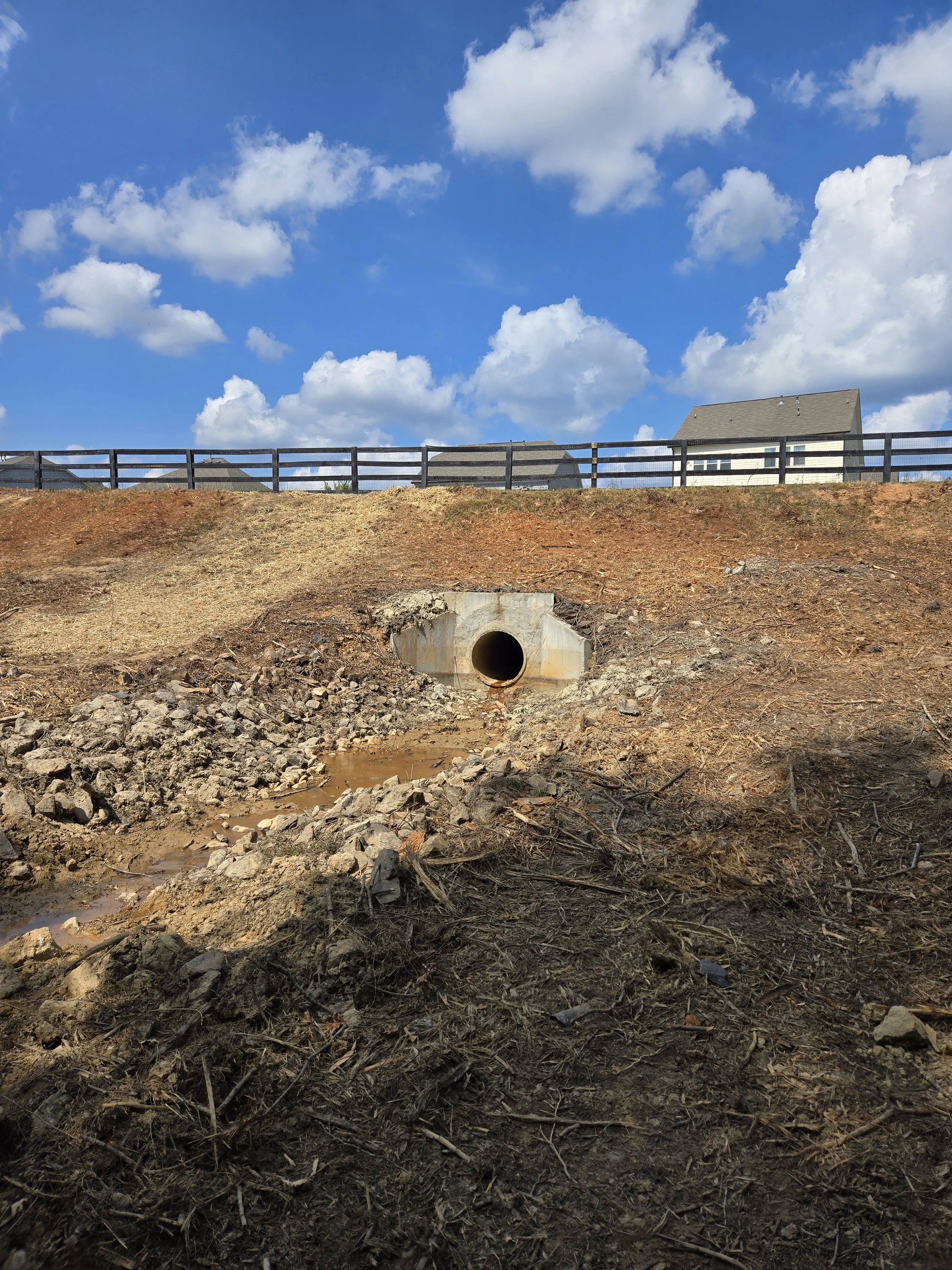 A yard with dry dirt and small rocks, featuring a large round drainage pipe at the bottom of the slope. The background shows a fence and houses under a partly cloudy blue sky.