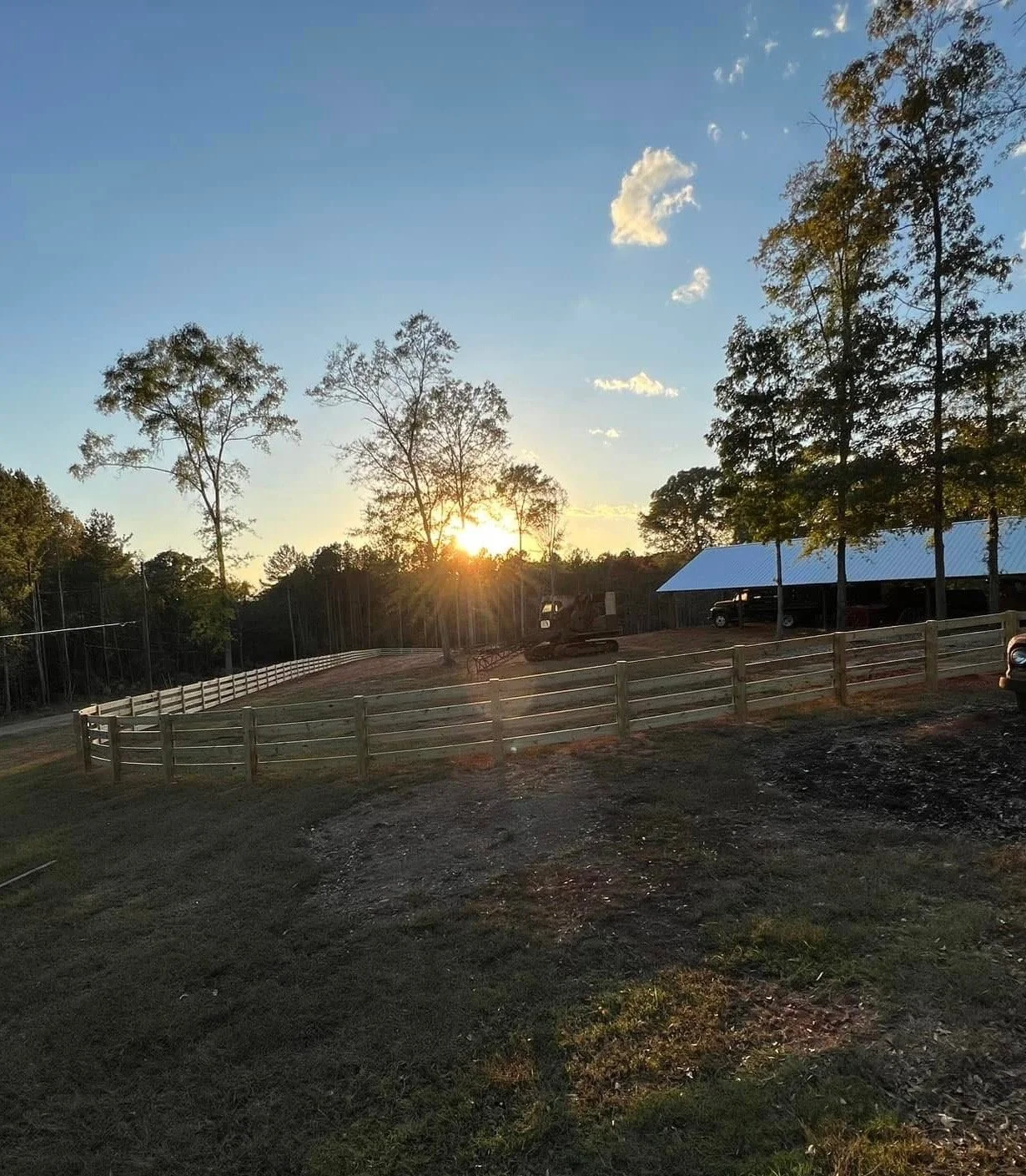 Sunset scene with trees, a fenced area, a carport, and vehicles, under a blue sky with clouds.