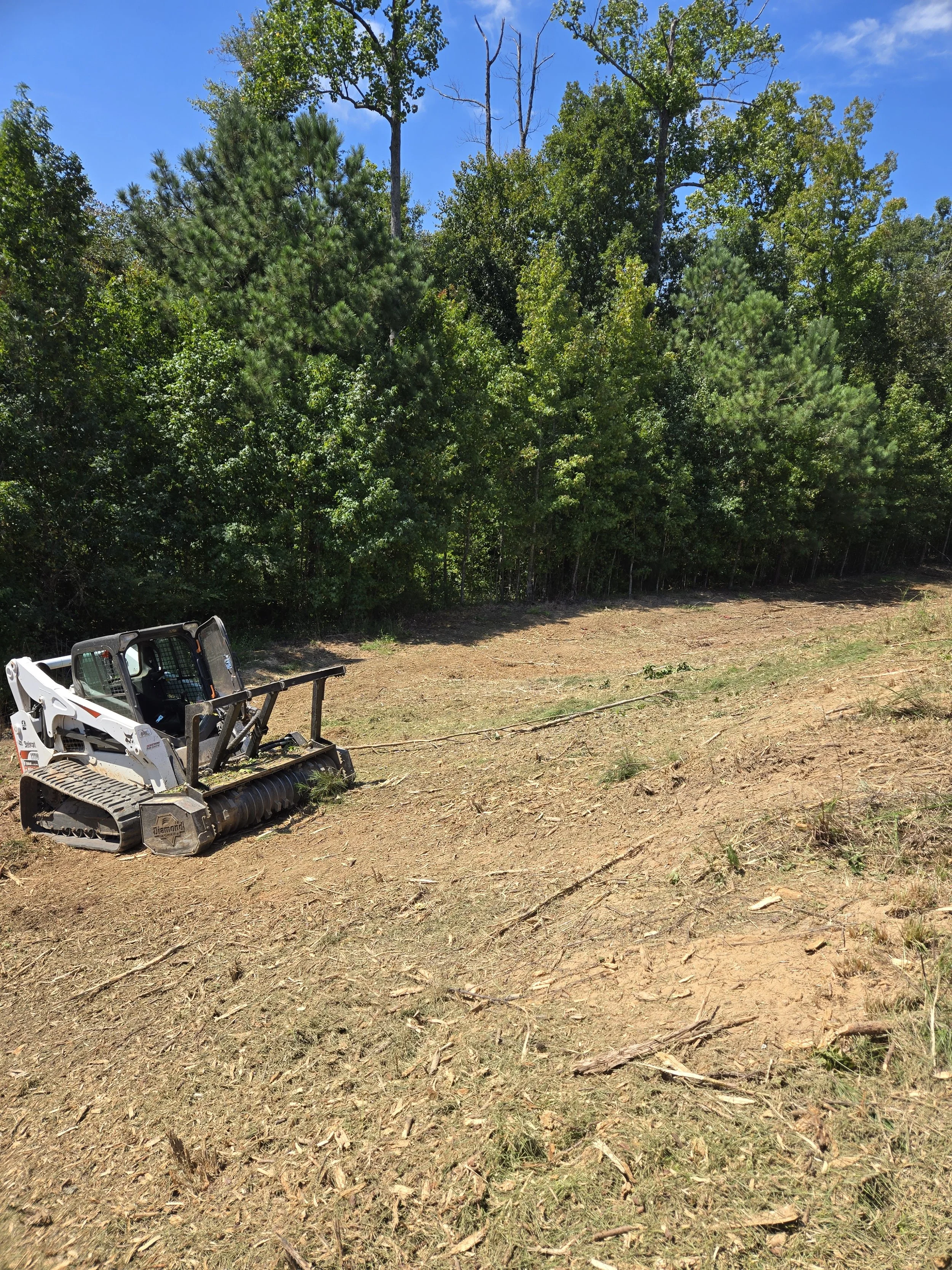 A small bulldozer parked on a dirt clearing, with a forested area of trees in the background under a bright blue sky.
