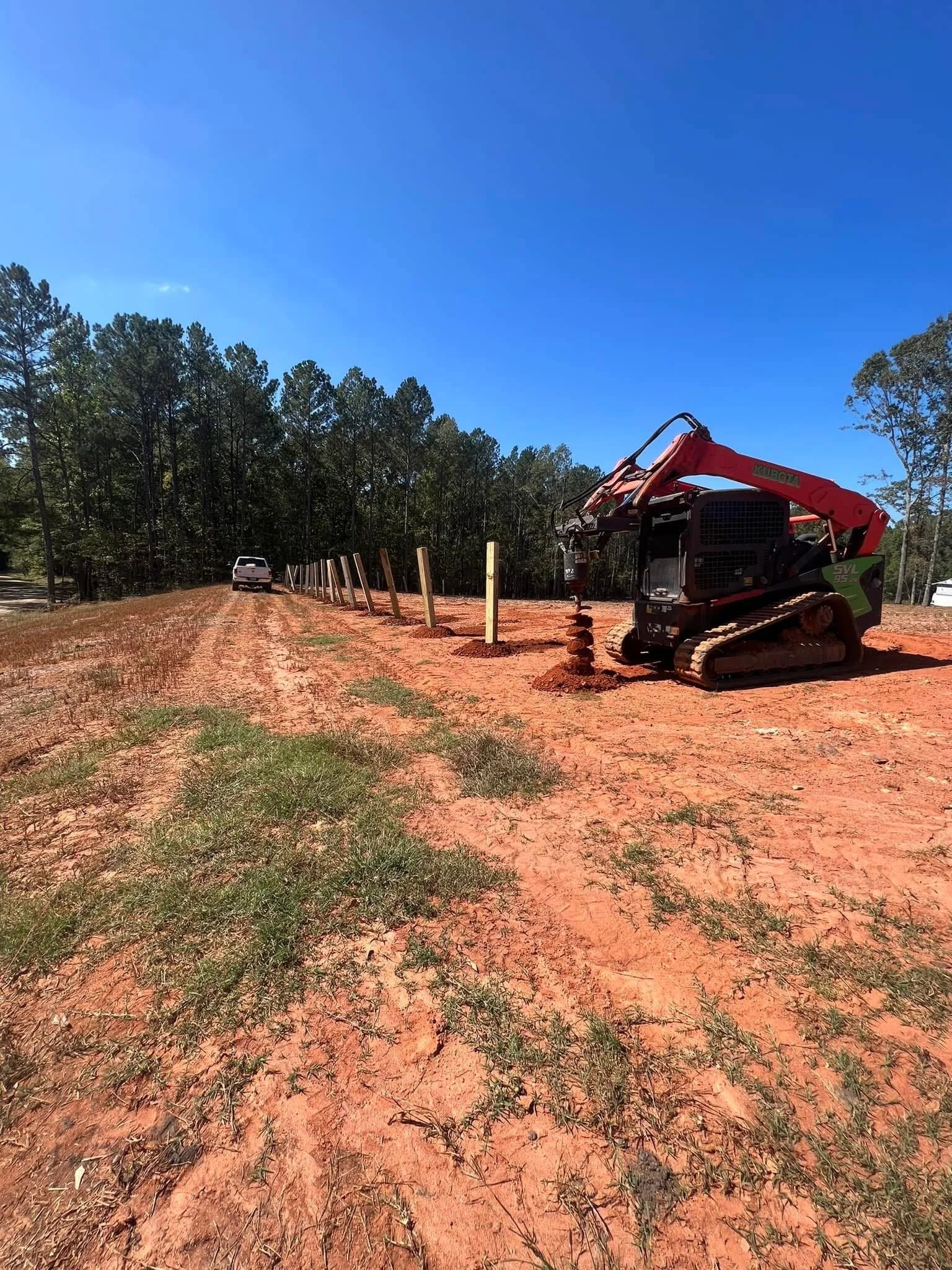 Construction site with a small red tracked excavator working on a dirt path, planting wooden posts along a boundary, near a wooded area under a clear blue sky.
