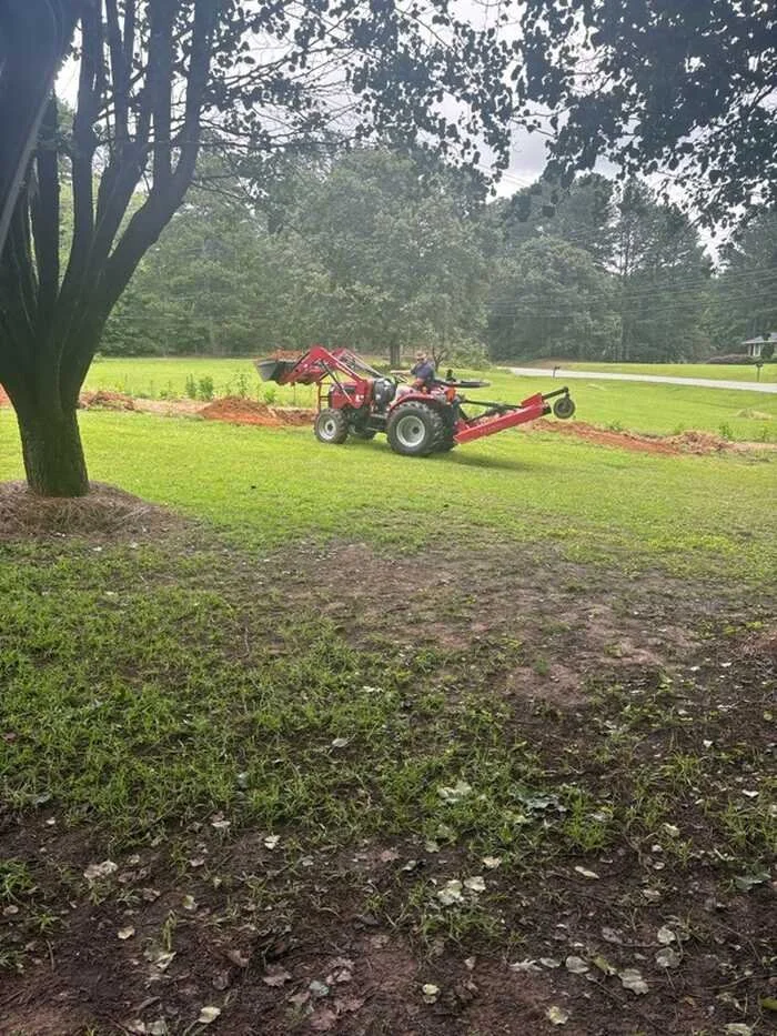 A red tractor on a grassy lawn, with some dirt patches and a large tree in the foreground, and a road and trees in the background.