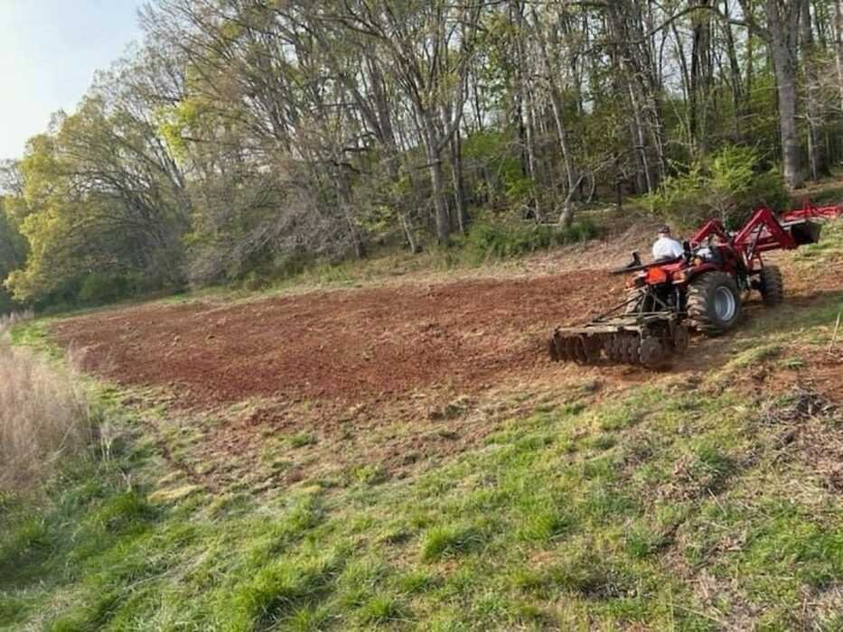 A person operating a red tractor tilling a field near a wooded area.