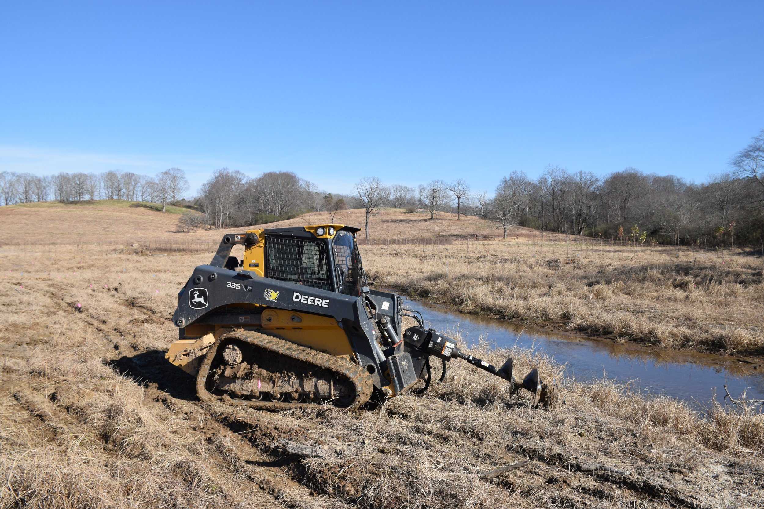 A John Deere skid steer loader working in a grassy field near a small water body, with bare trees and a clear blue sky in the background.