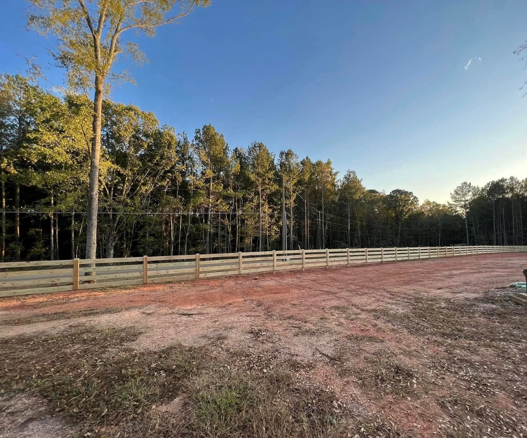 Construction site with a dirt road, new wooden fence, and trees in the background under a clear blue sky.