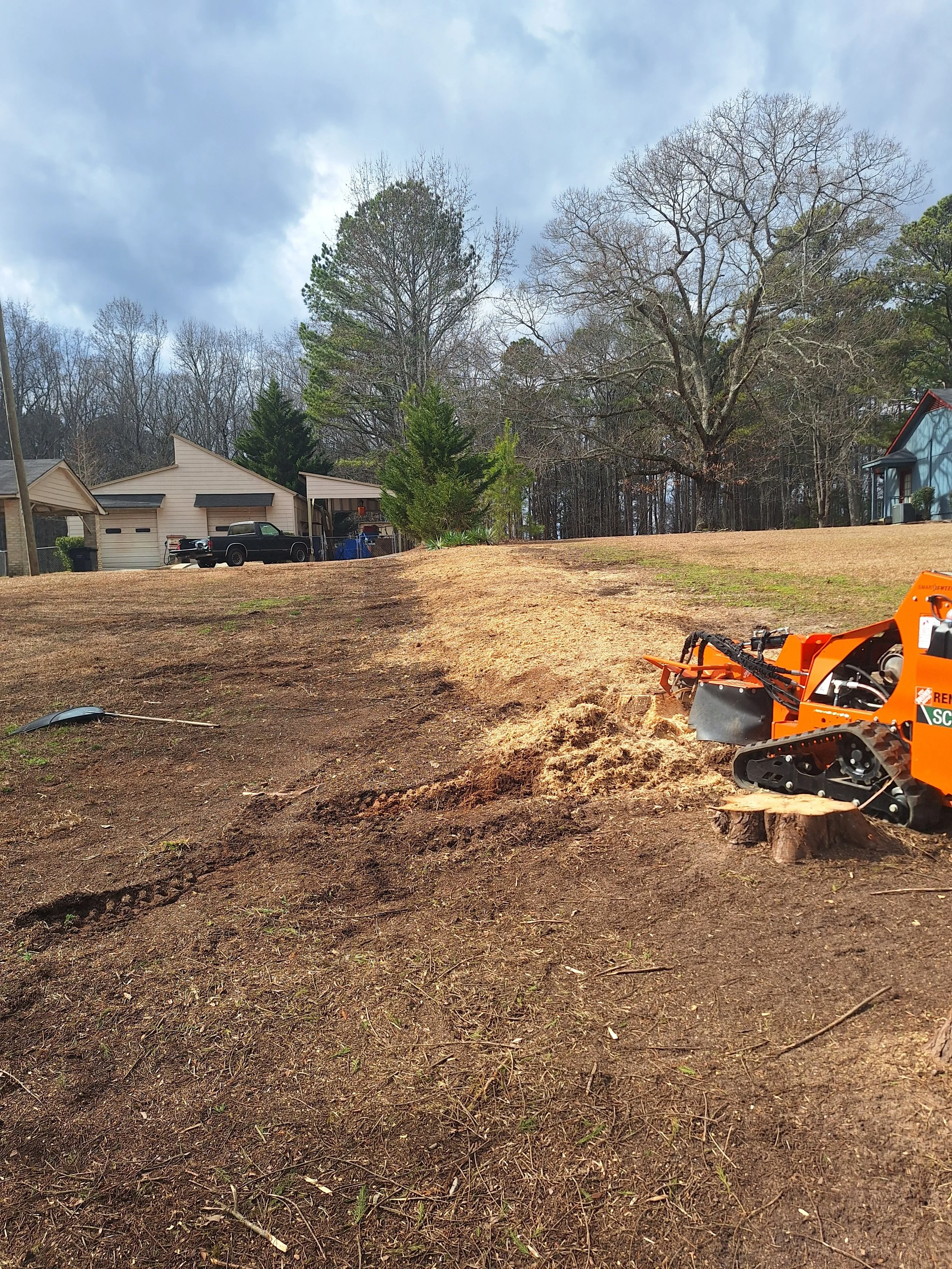 A small orange tracked loader working on clearing and leveling a sloped dirt yard with a tree stump in the foreground. Residential houses and trees are visible in the background under a partly cloudy sky.