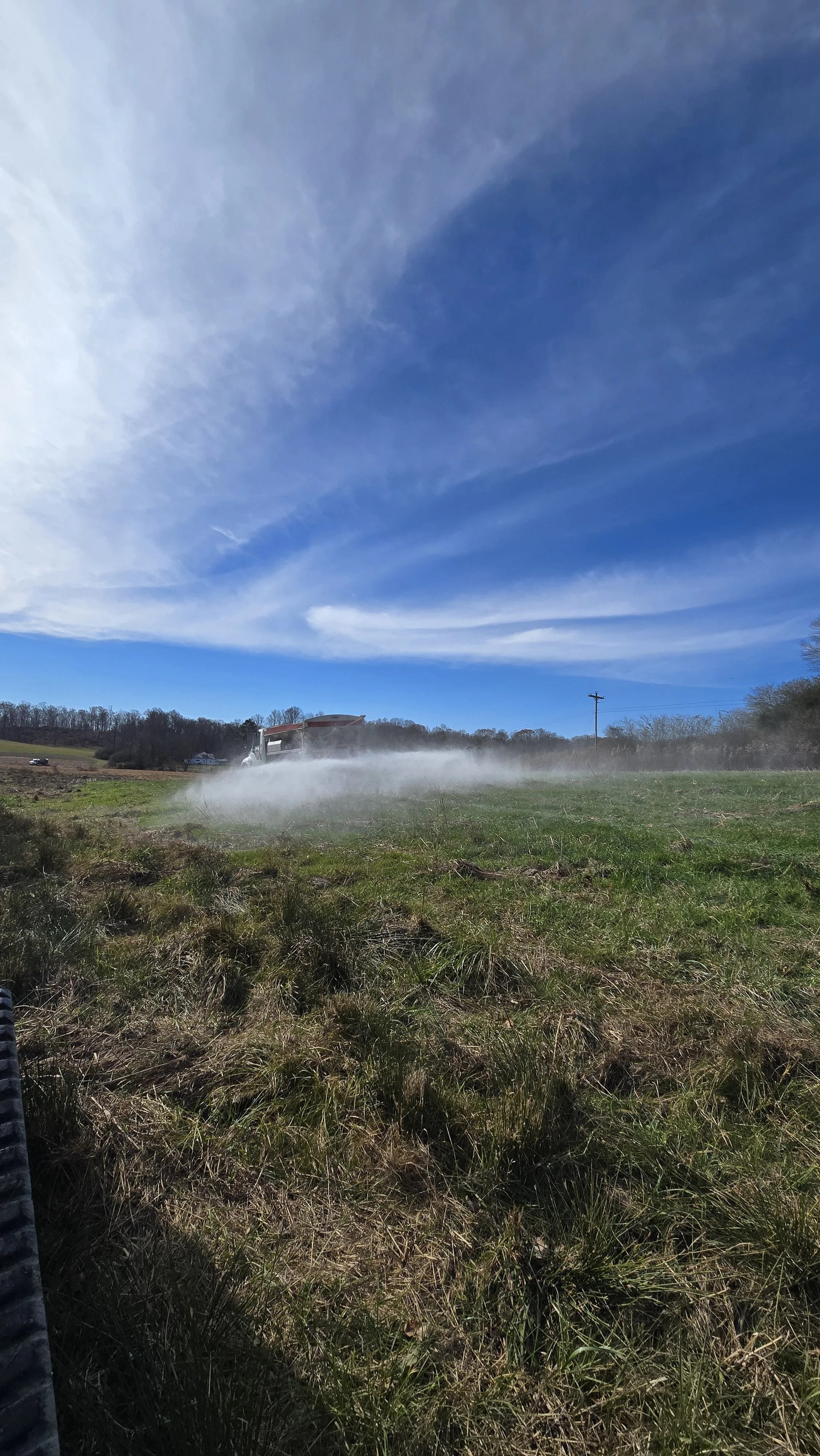 A blue sky with some clouds over a grassy field where a tractor is spraying or watering the field, creating a mist or spray visible in the air.