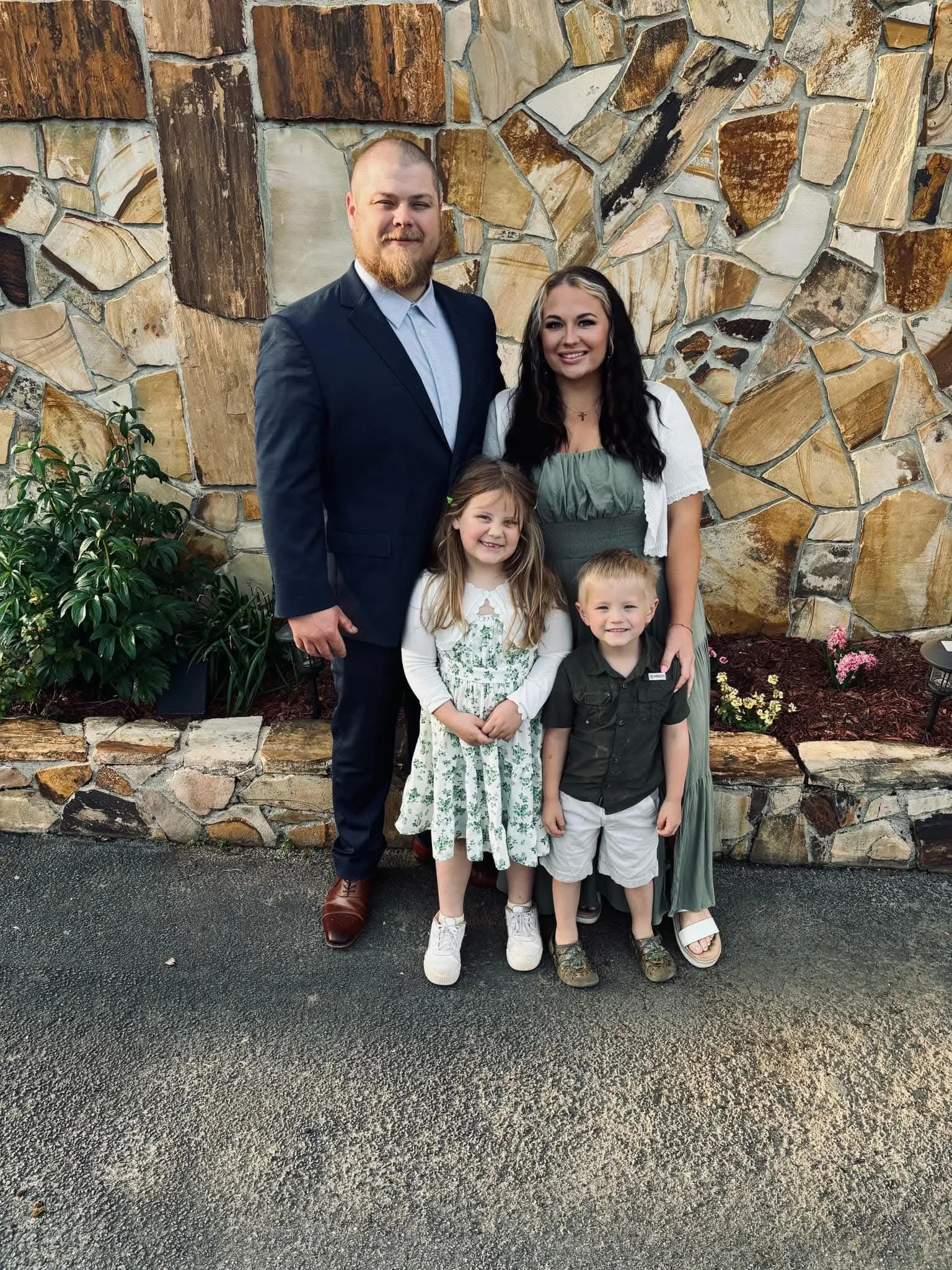 A family of five standing outdoors in front of a stone wall, smiling for the camera. The father is wearing a navy suit, and the mother has long dark wavy hair, wearing a green dress with a white jacket. The two children, a girl with long blonde hair in a white dress with green floral pattern and white sneakers, and a boy with short blonde hair in a green shirt and white shorts, are standing in front of their parents.