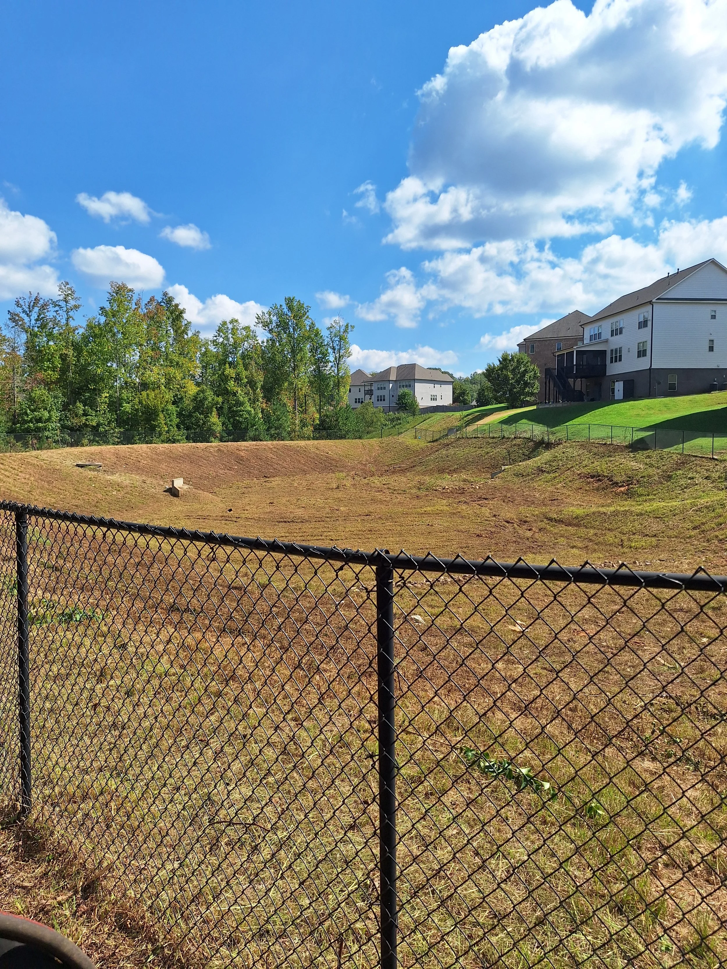 Empty yard with a chain-link fence, grassy slopes, and residential houses under a partly cloudy sky.