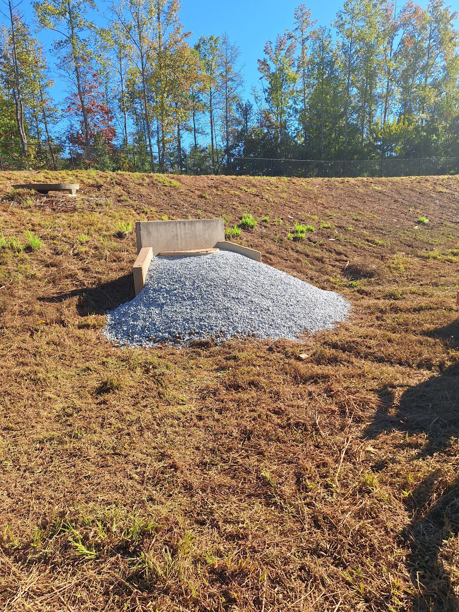 Construction site on a hill with a pile of gravel in a forming area, bordered by small concrete blocks, with a background of trees and a bright blue sky.