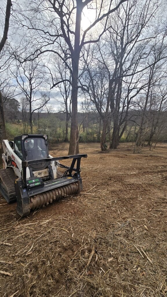 A construction or landscaping site with a compact skid steer loader equipped with a tiller attachment, parked on freshly tilled soil with leafless trees and a cloudy sky in the background.