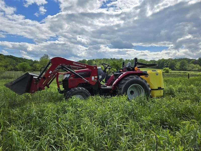 Red and yellow Mahindra tractor with front loader attachment working in a green field under cloudy sky