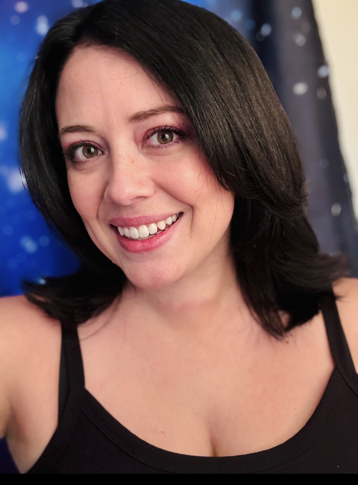 A woman with dark, shoulder-length hair, smiling with white teeth, wearing a black top, and posing for a selfie against a blue background with white spots.
