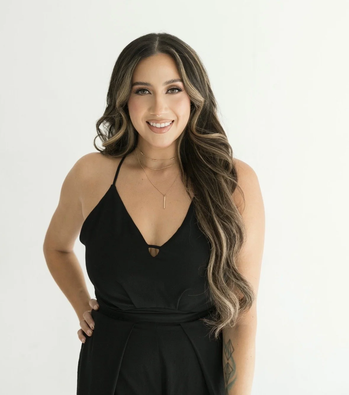 A smiling woman with long wavy hair wearing a black sleeveless dress and layered necklaces, standing against a plain light-colored background.