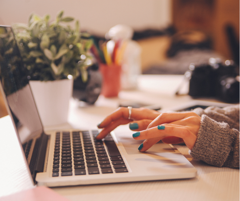 Person typing on a laptop with a plant, pen holder, and camera on a cluttered desk.