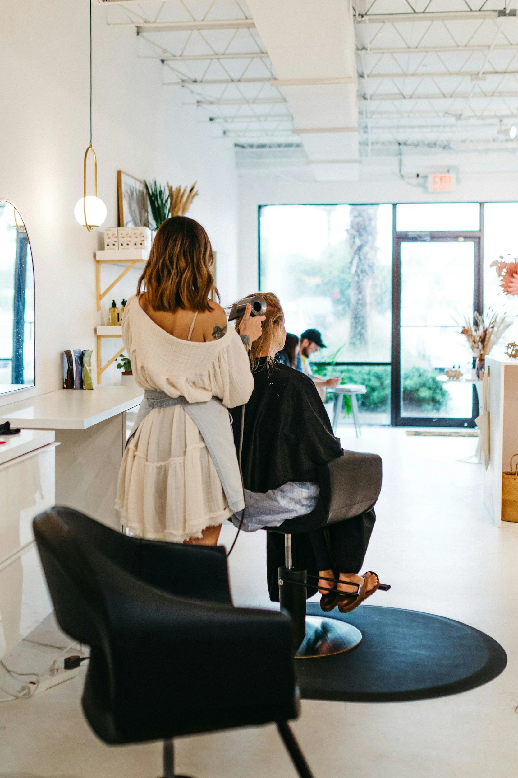 A woman getting a haircut at a modern salon with white walls and large windows, seated in a black salon chair, while a hairdresser dries her hair with a hand-held blow dryer.