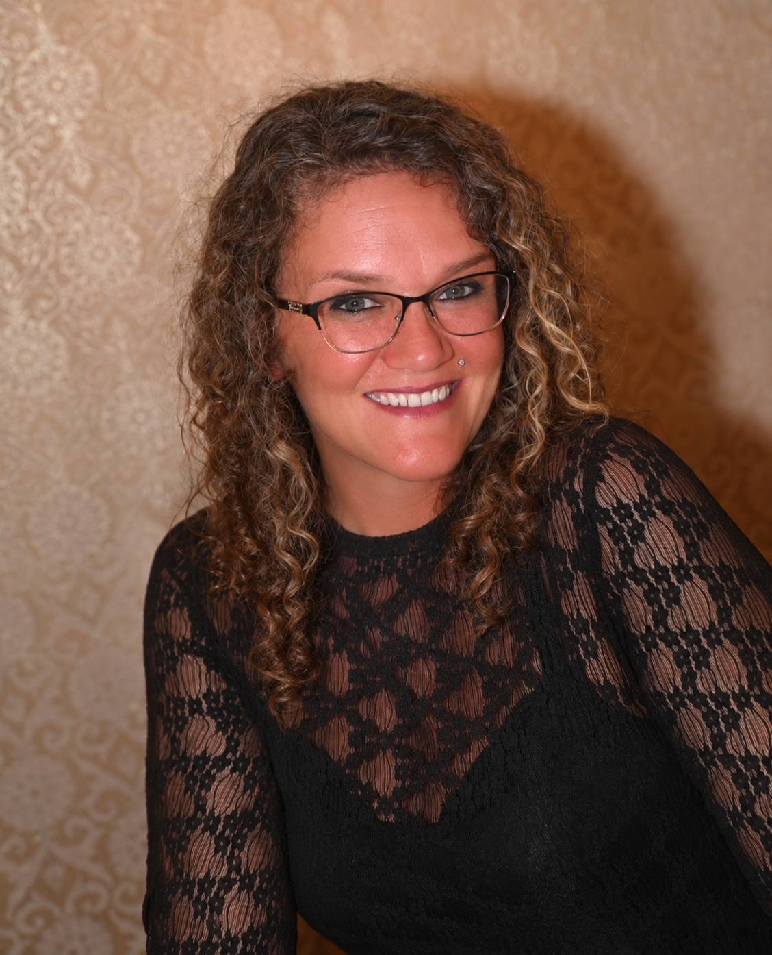 A woman with curly hair, glasses, and a nose piercing smiling, wearing a black lace top, against a gold-patterned background.