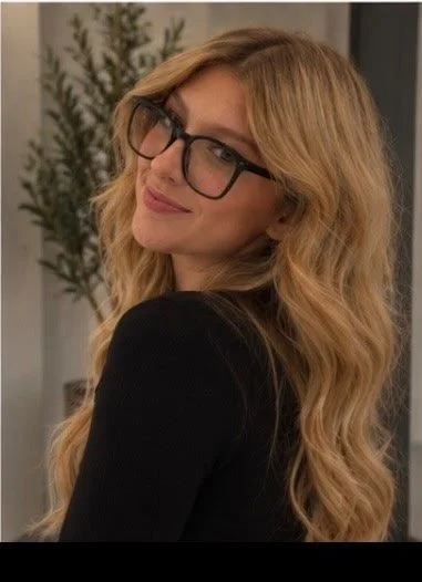 A woman with long, wavy blonde hair and black glasses, smiling and looking over her shoulder in a casual indoor setting with a plant in the background.