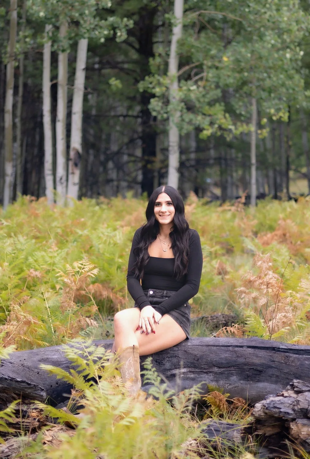 A woman with long black wavy hair, dressed in black, sitting on a fallen log in a forest meadow with ferns and trees in the background.