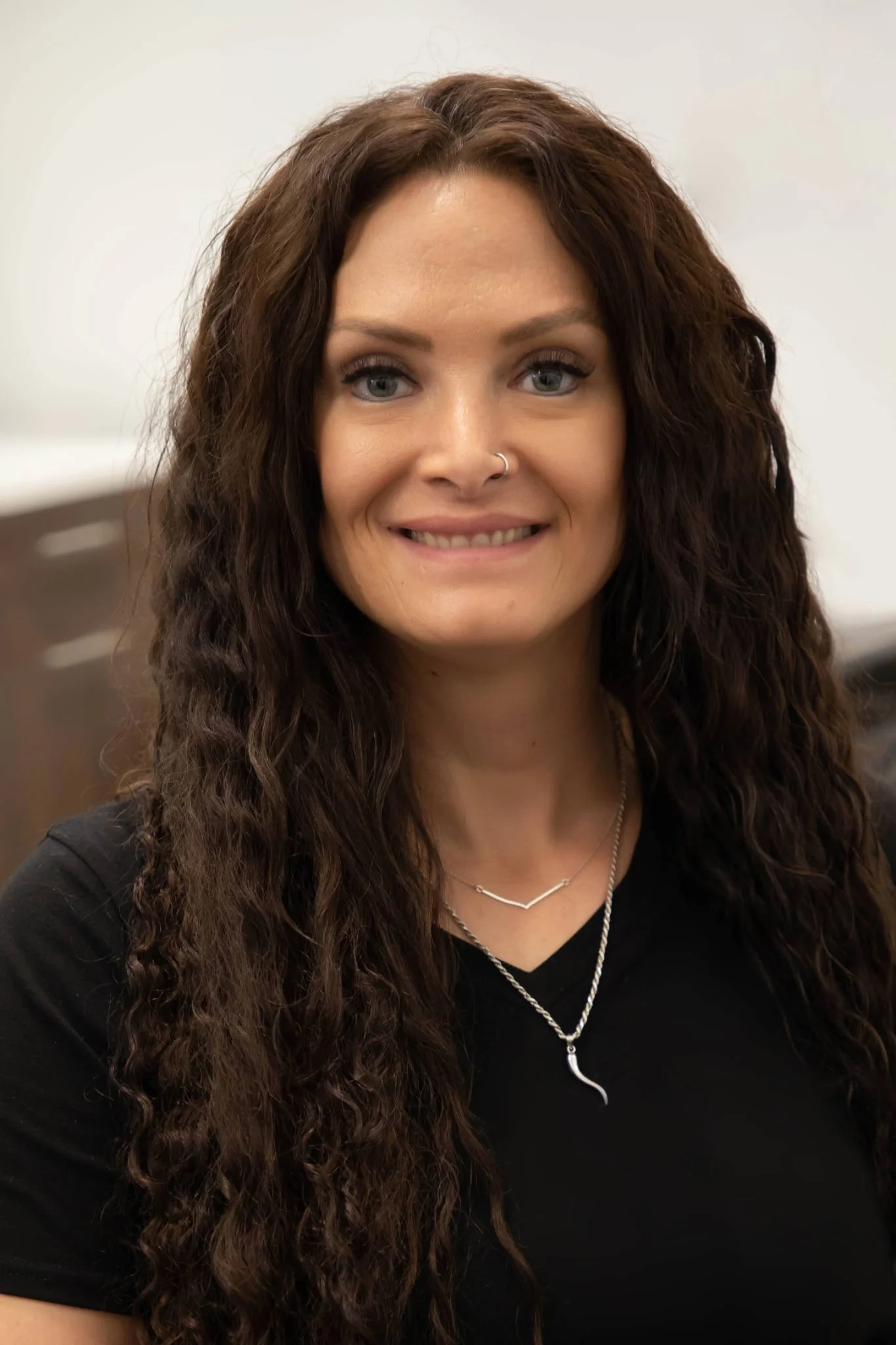 Close-up portrait of a woman with long, curly brown hair and light blue eyes, smiling, wearing a black top and layered silver necklaces, against a white background.