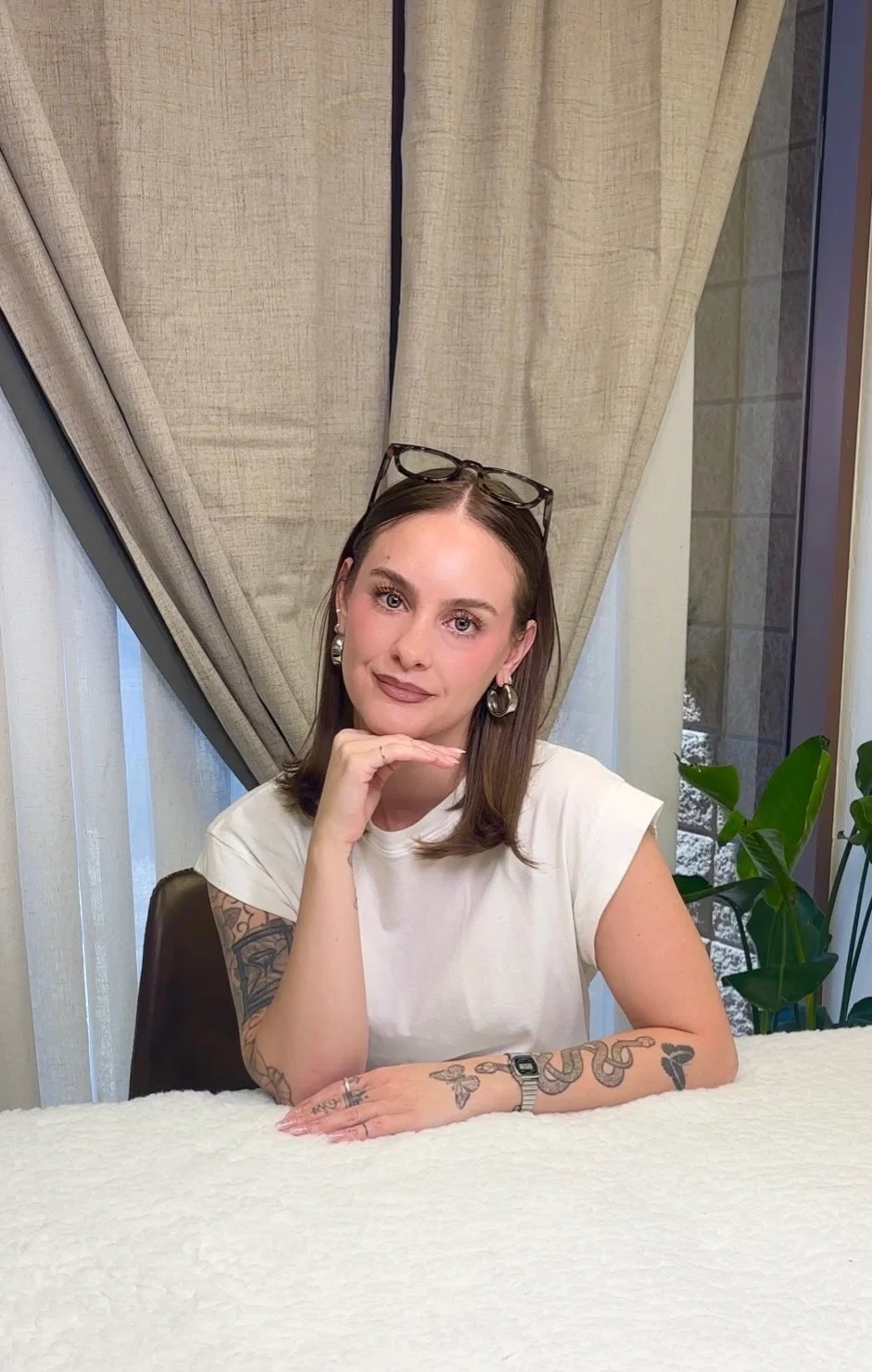 A woman with shoulder-length brown hair, wearing glasses on her head, silver earrings, and a watch, sitting at a table with her chin resting on her hand, smiling at the camera. She has tattoos on her arms, and a beige curtain and a plant are visible in the background.