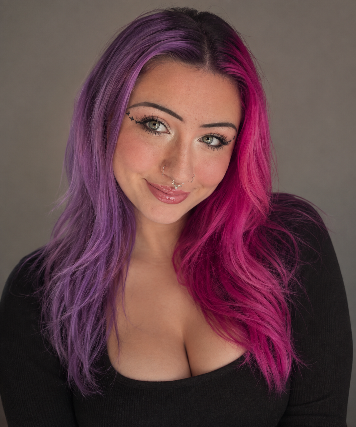 A woman with vibrant, split-colored hair, purple on the left and pink on the right, smiling at the camera against a neutral background.