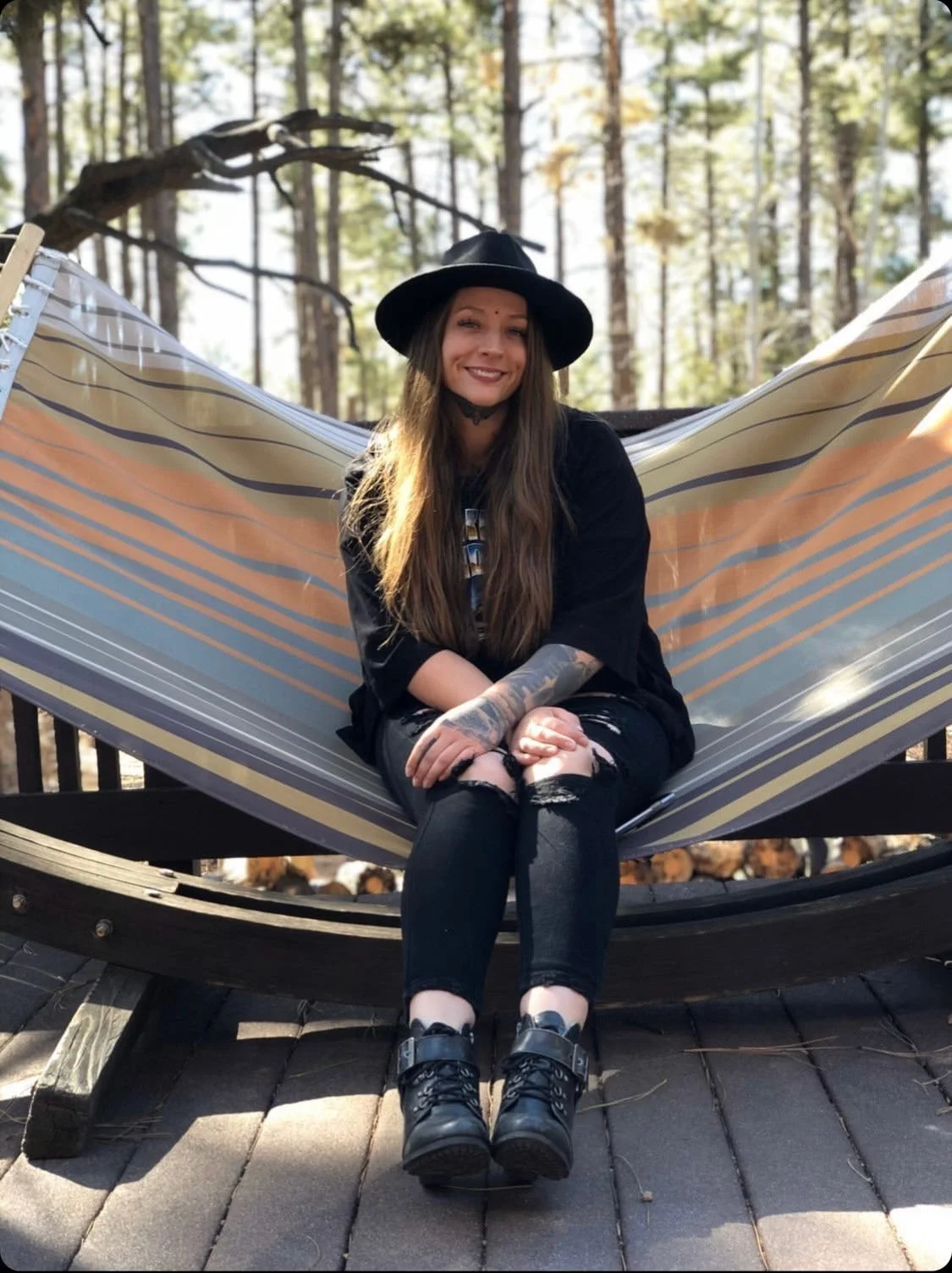 Young woman sitting in a hammock outdoors in a wooded area, wearing a black hat, black distressed clothing, and smiling.