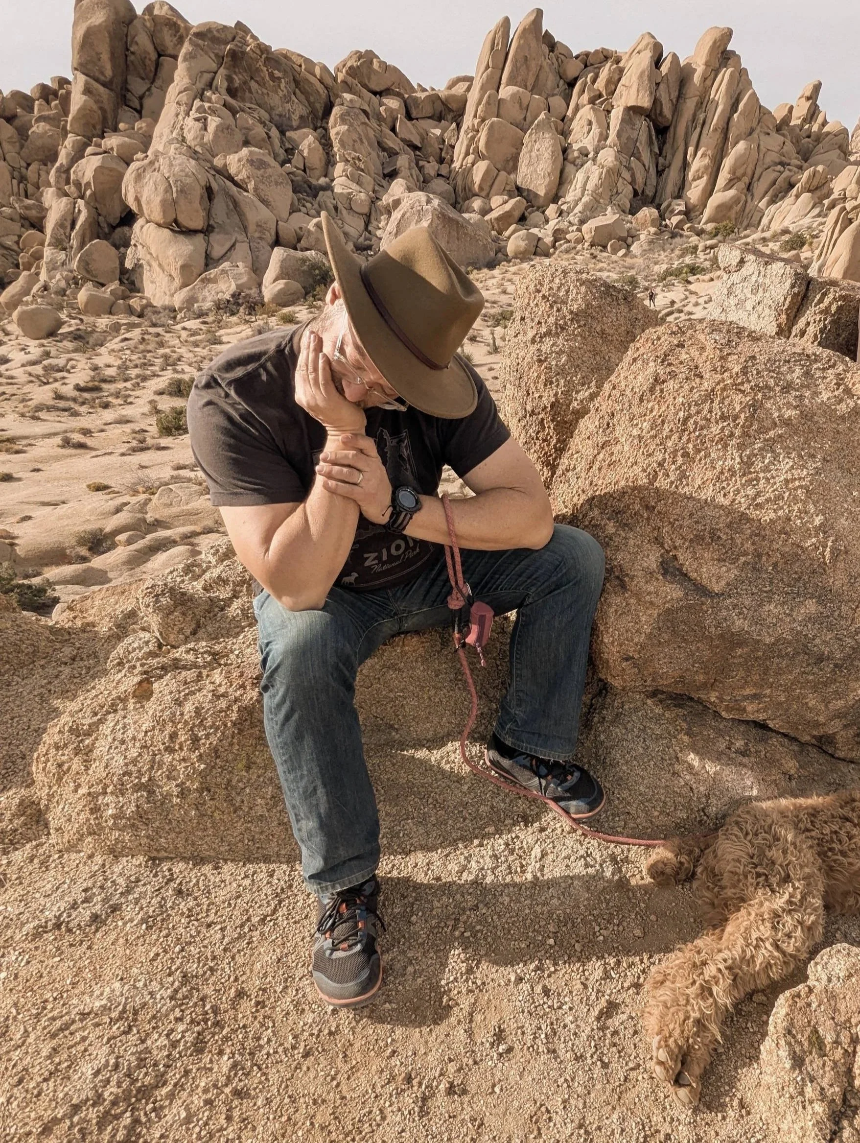 A man in a black T-shirt, jeans, and athletic shoes with a tan hat, sits on a large rock in a desert landscape with large boulders and rocky formations in the background. He appears to be resting on a hike, with his head tilted down and hand supporting his chin. A small curly-haired dog lies on the ground nearby on a leash.