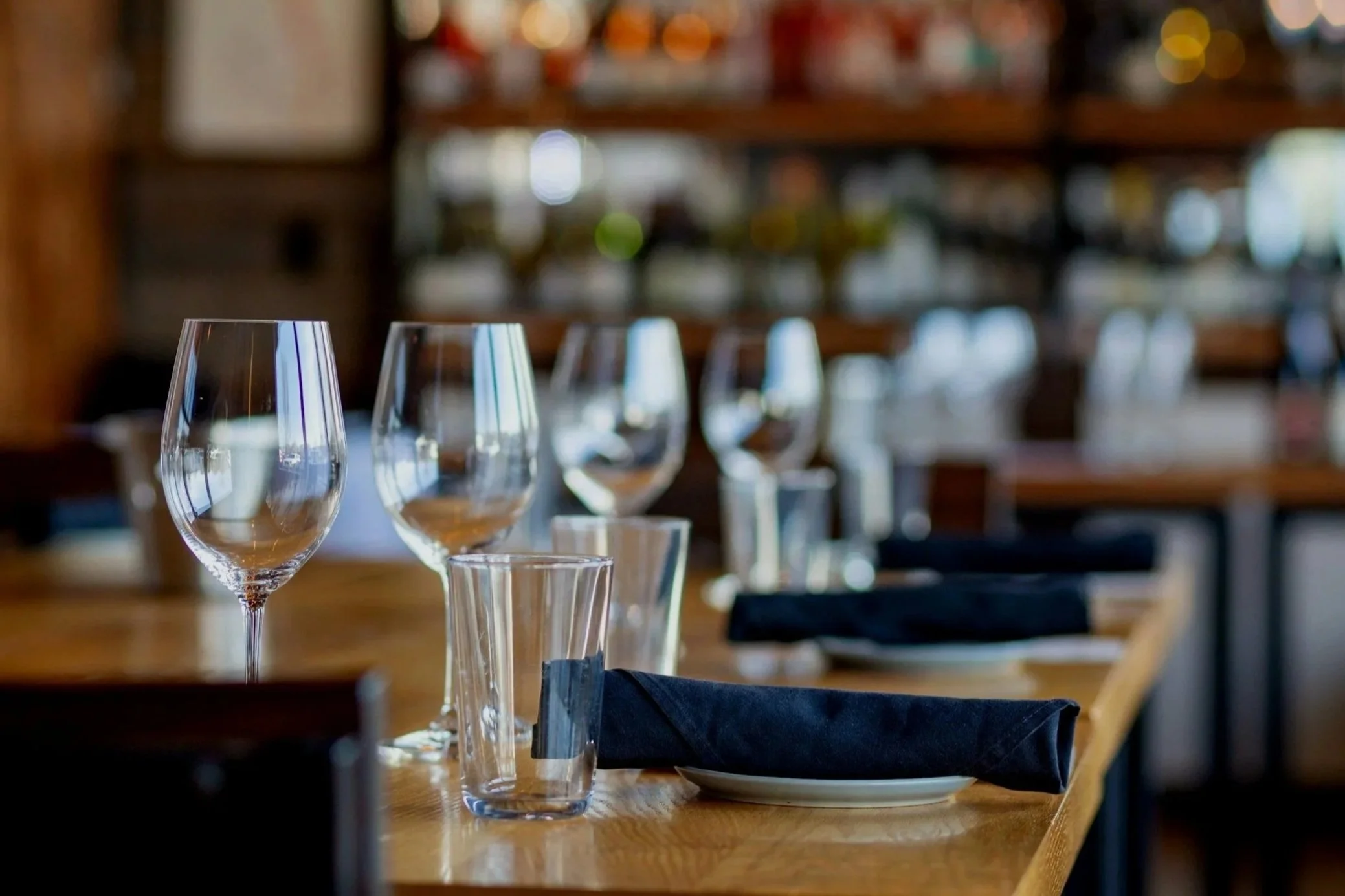 Empty wine glasses, water glasses, and black cloth napkins on a wooden table in a restaurant or bar setting with blurred background.