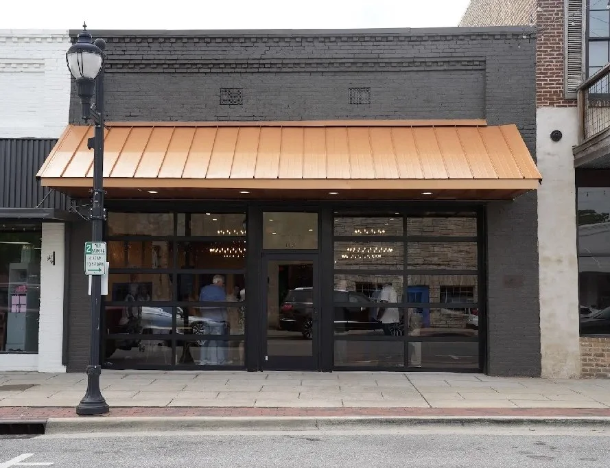 A storefront with large glass windows and a black door, with an orange metal awning above. A streetlamp and parking sign are in front, and reflections of cars and people are visible in the windows.