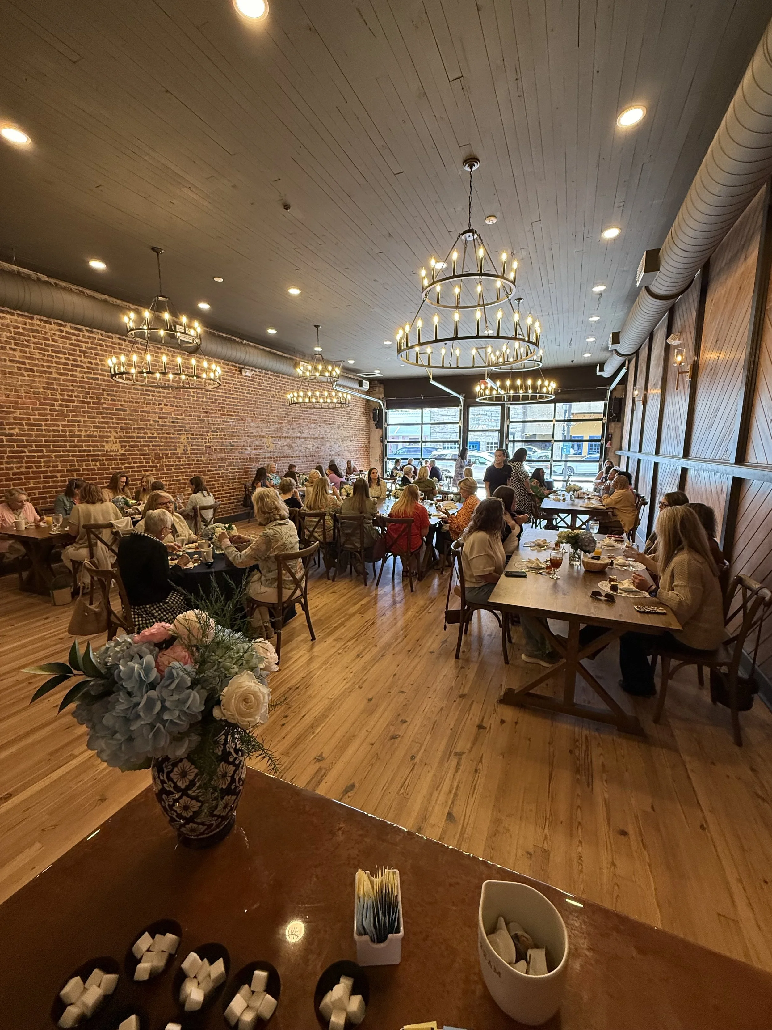 Interior of a restaurant with exposed brick walls and wooden flooring. Several large chandeliers hang from the ceiling, and patrons are seated at tables along the walls and in the center. The foreground shows a counter with a floral vase, sugar packets, and condiment containers.