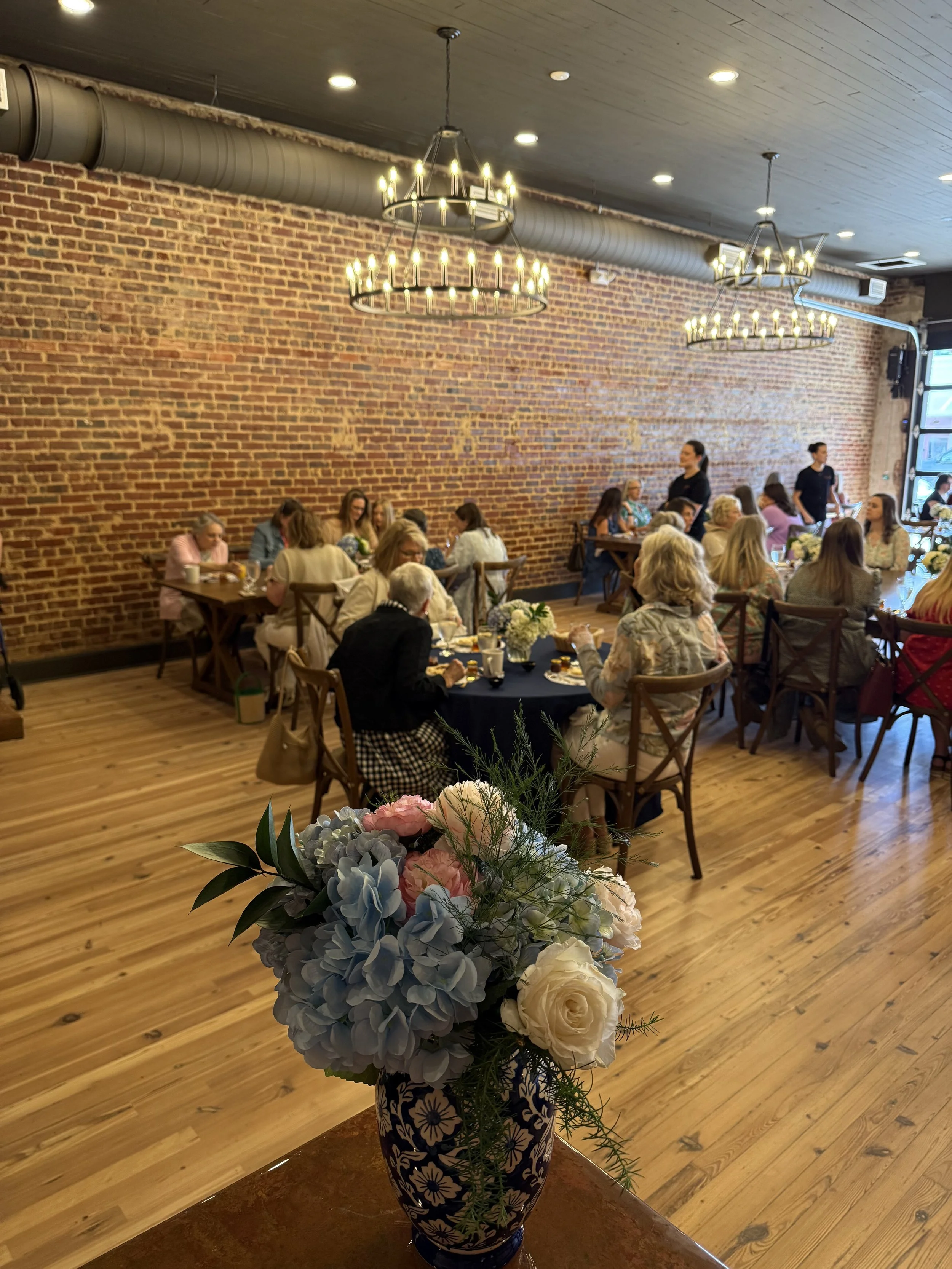 A large floral arrangement in a blue and white patterned vase, placed on a wooden table in a restaurant or event space, featuring white roses, pink hydrangeas, blue hydrangeas, and green foliage.