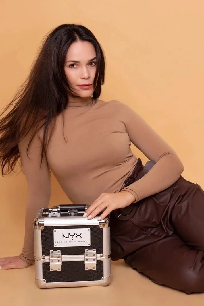 A woman with long brown hair posing against a light brown background, wearing a tan long-sleeve turtleneck and dark brown pants, with a makeup case labeled NYX on the ground.