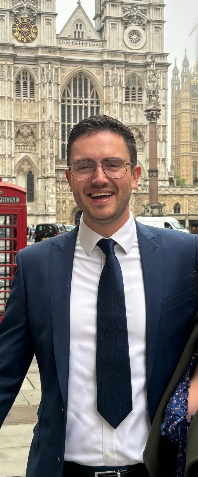 A man in a blue suit and tie smiling outdoors in front of a historical Gothic-style building with intricate architectural details, large stained glass windows, and clock towers.