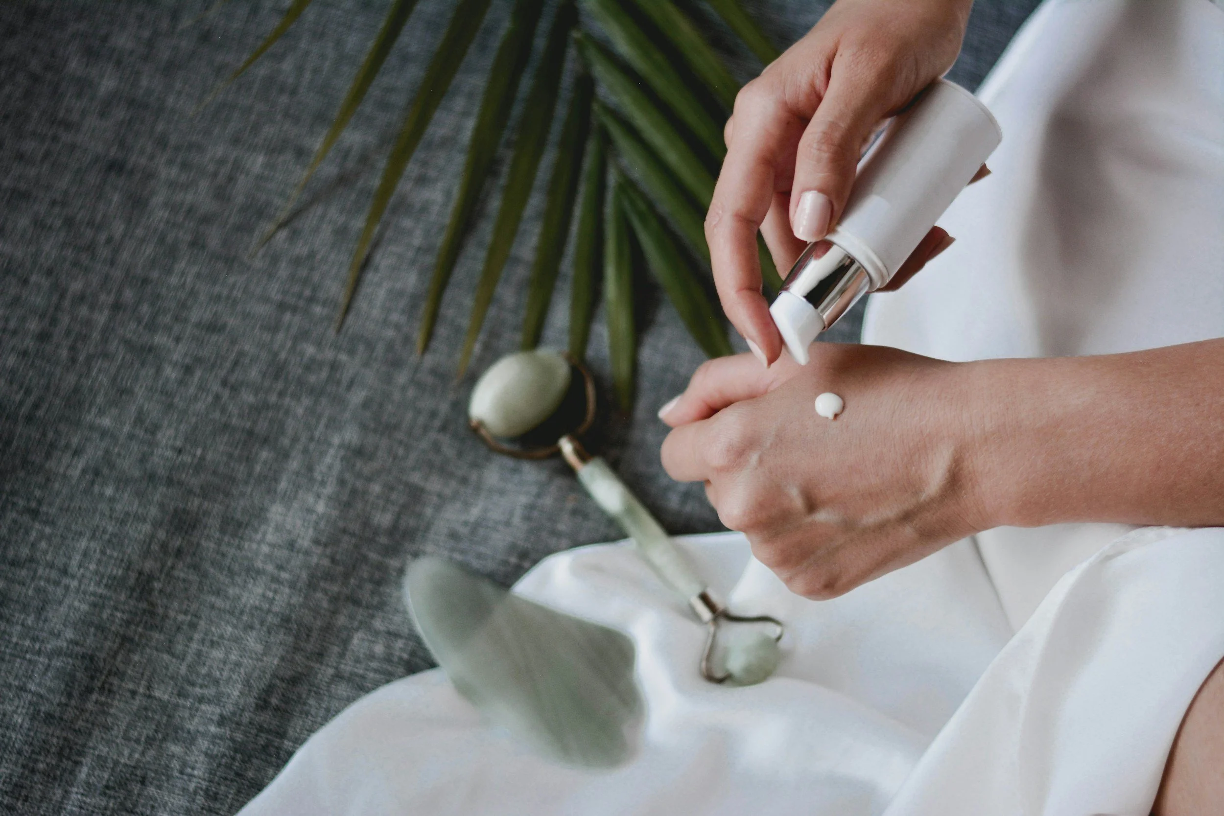 A person applying lotion or cream from a pump bottle onto their hand. There is a jade facial roller and a green plant leaf on a gray textured surface.