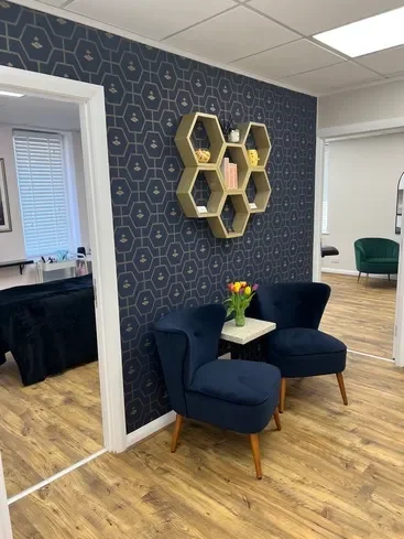 Corner of a room with two navy blue armchairs and a small white table with a vase of yellow and pink tulips on it, against a dark blue patterned wall with honeycomb-shaped shelves hanging on it, and a window with blinds to the left.