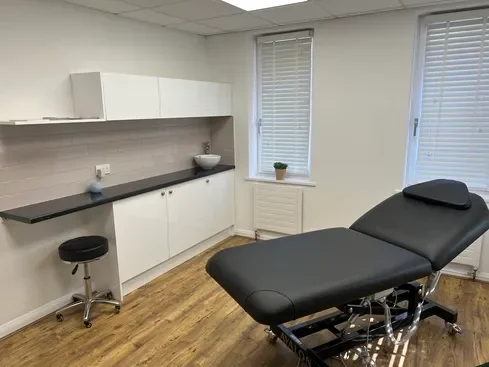 Medical examination room with examination table, stool, cabinets, and windows.