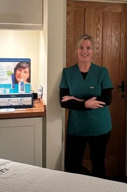 A woman in a teal medical uniform smiling with arms crossed, standing next to a wooden door in a room with a bed and a promotional display for skincare products.