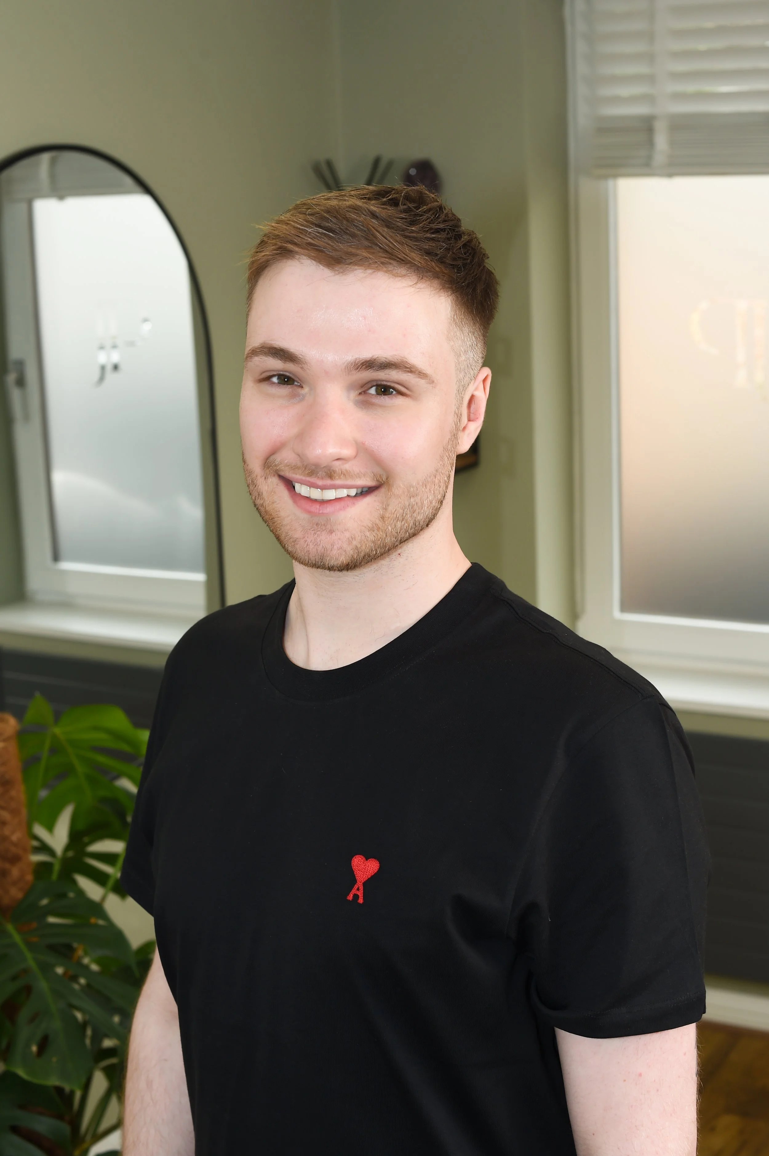 Young man with light brown hair and beard smiling, wearing a black t-shirt with a small red heart and knife logo on the chest, standing indoors near a window.