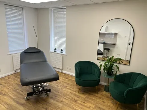 A medical examination room with a black examination chair, two green velvet chairs, a tall mirror, a small black side table with a plant, and a wall with two windows.