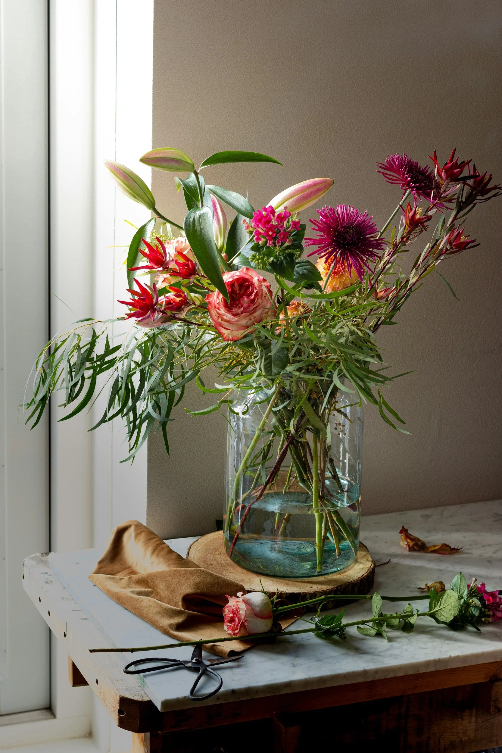 A bouquet of pink and purple flowers in a clear glass vase on a marble table near a window.