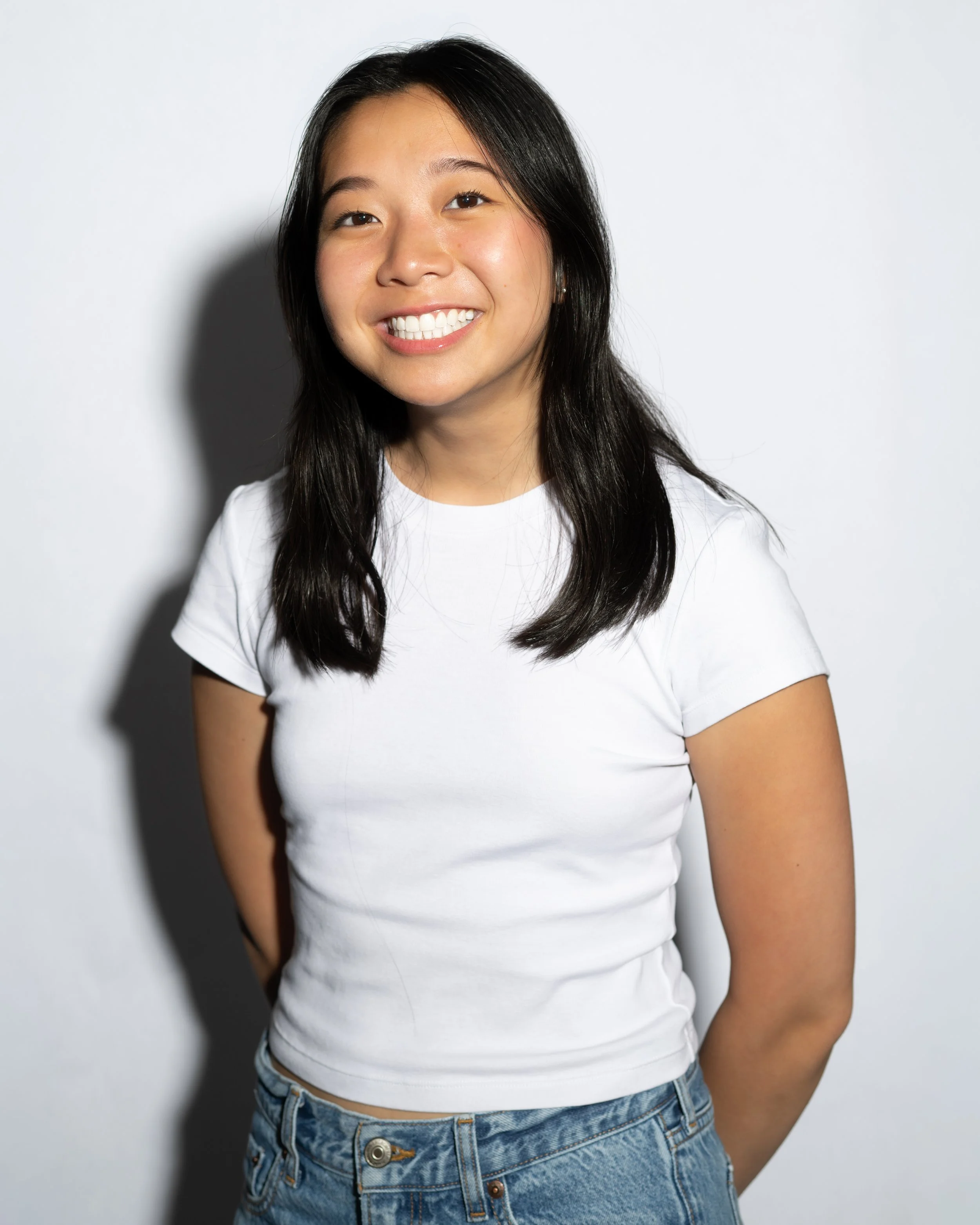 Young woman with long black hair, smiling, wearing a white t-shirt and blue jeans, standing against a plain white background.
