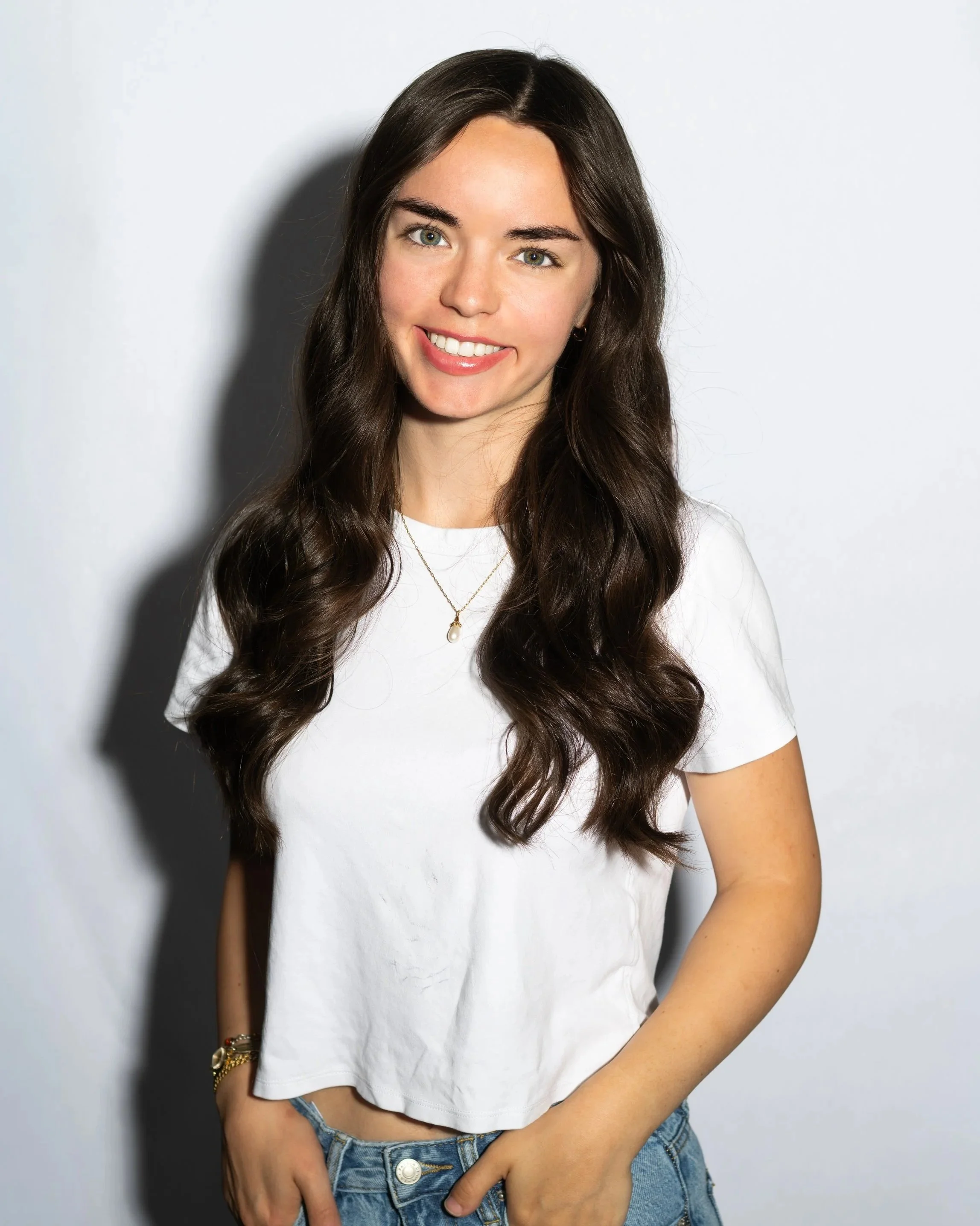 Young woman with long wavy brown hair smiling, wearing a white t-shirt and blue jeans, standing against a plain white background.
