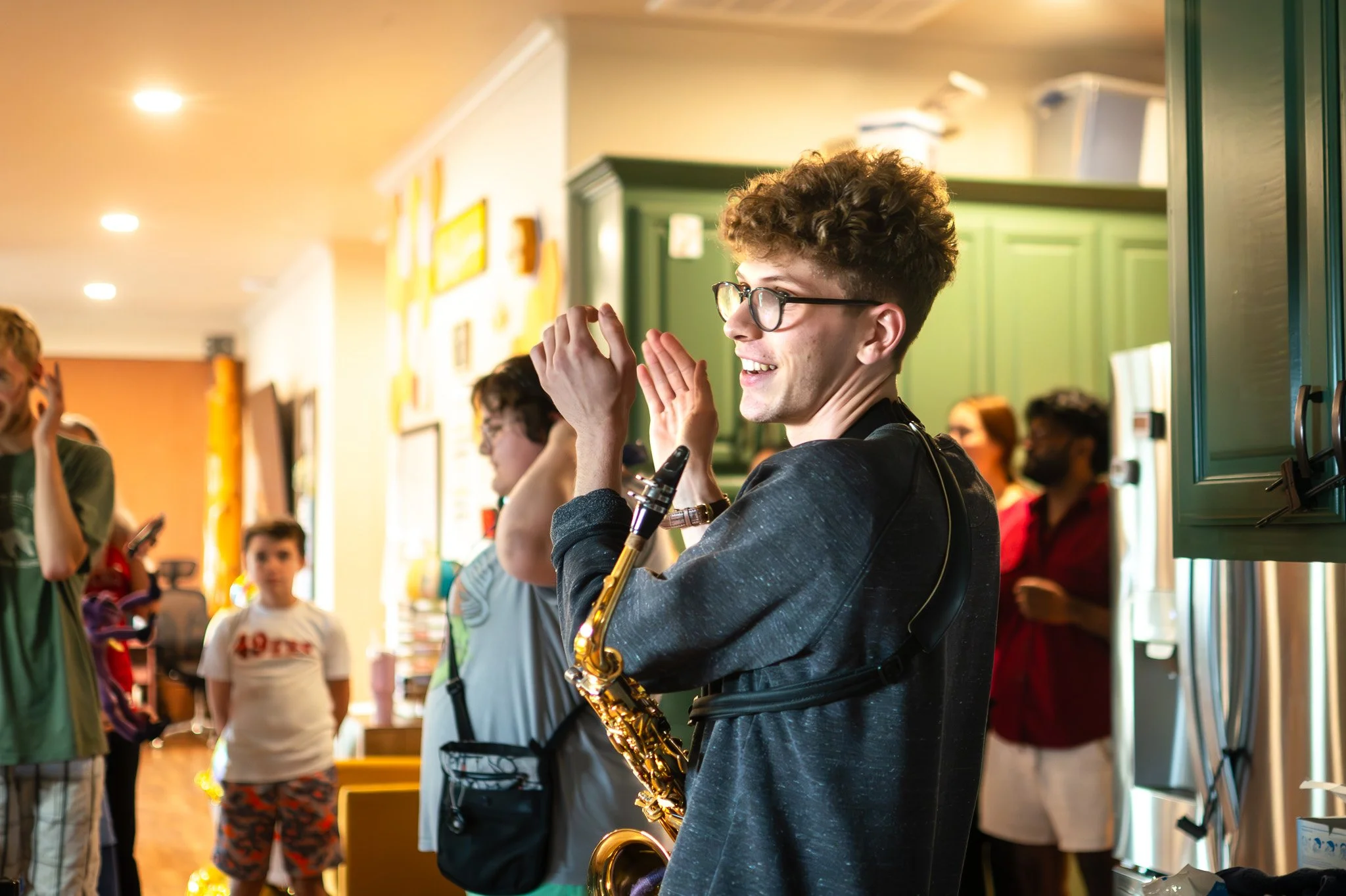 Young man with curly hair and glasses playing saxophone in a social gathering indoors, surrounded by people and colorful decor.