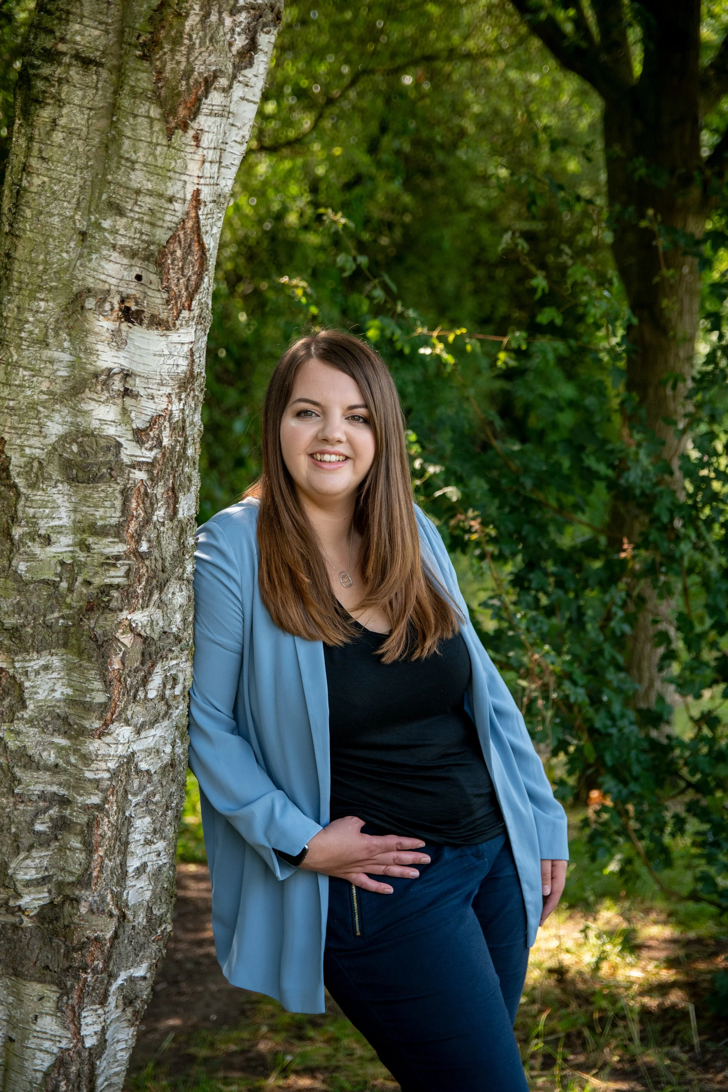A woman with long brown hair standing outdoors next to a tree, smiling at the camera, surrounded by green foliage.