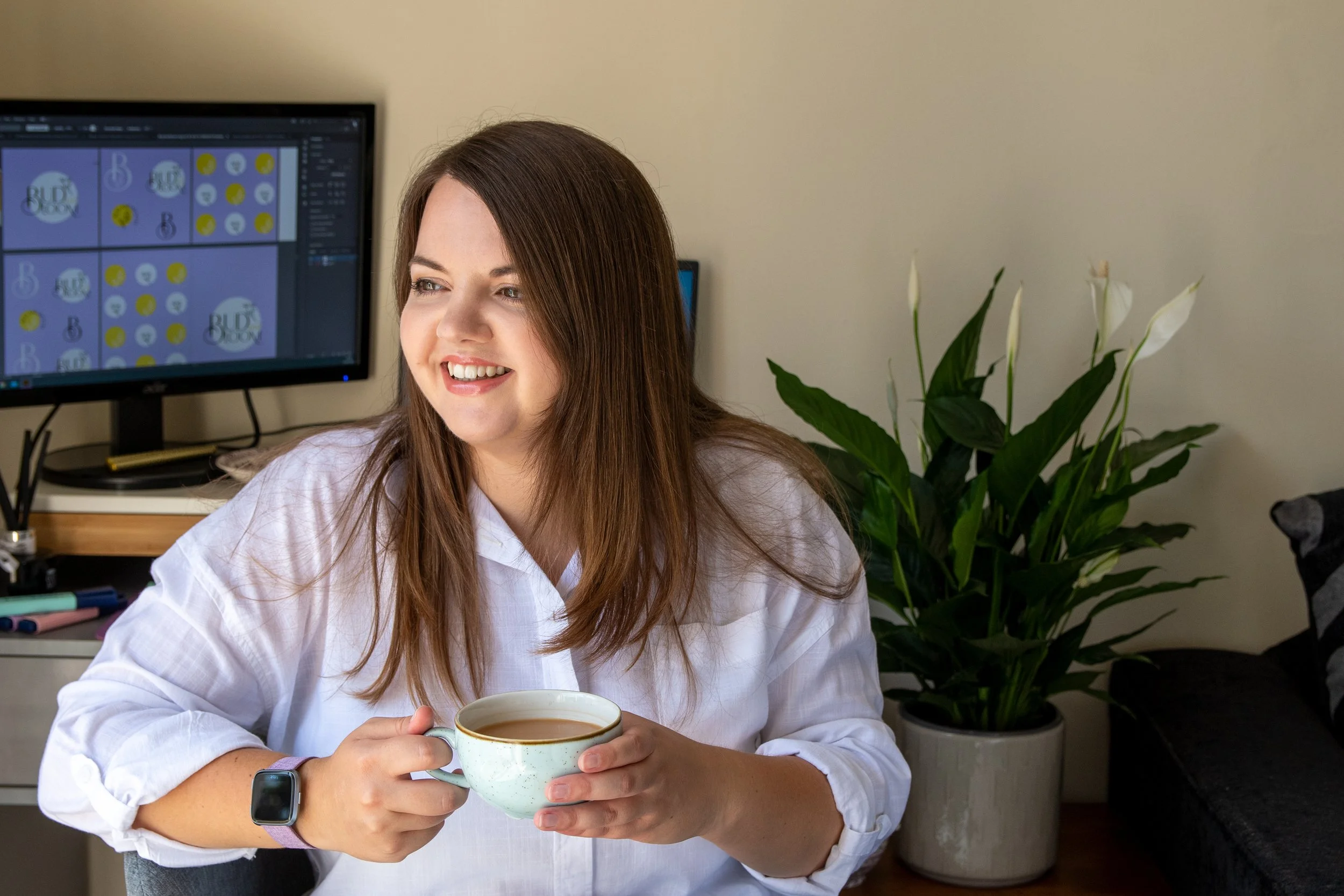 A woman with brown hair sitting at a desk, holding a mug of coffee, with a computer monitor and a potted plant in the background.